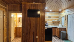 A wooden interior with a kitchen area, a mounted television, and a trash bin. - “The Bear Cave” WhittleSticks Cabin in Lancing TN (Lancing)