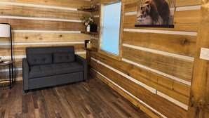 A cozy room with wooden walls, a gray sofa, a small table, and a window with blinds. - “The Bear Cave” WhittleSticks Cabin in Lancing TN (Lancing)