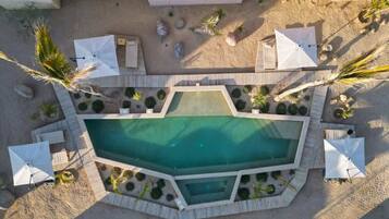 Vista aérea de una zona de piscina con una pileta rectangular, rodeada por una terraza de madera, sombrillas blancas y palmeras.