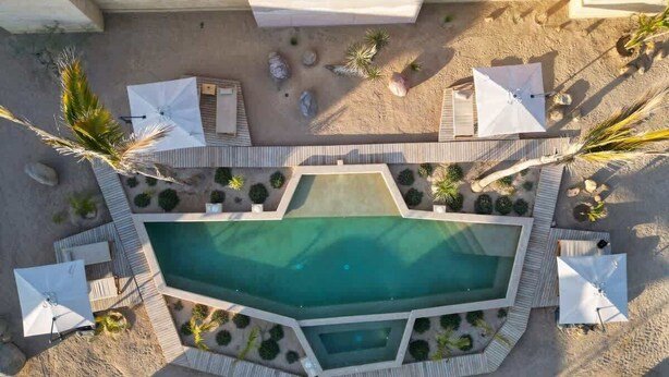 Vista aérea de una zona de piscina con una pileta rectangular, rodeada por una terraza de madera, sombrillas blancas y palmeras.