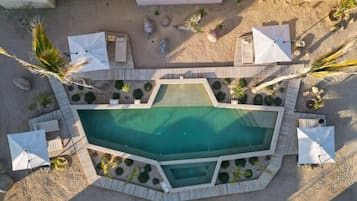 Vista aérea de una zona de piscina con una pileta rectangular, rodeada por una terraza de madera, sombrillas blancas y palmeras.