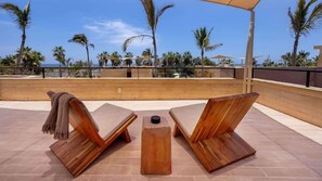 Eine Holz-Lounge mit zwei Sesseln und einem kleinen Tisch, mit Blick auf einen Strand mit Palmen und einem klaren Himmel.