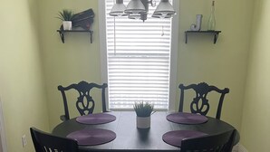 A dining area with a round table, black chairs, and a chandelier. There are two shelves with decorative items and a window with blinds.