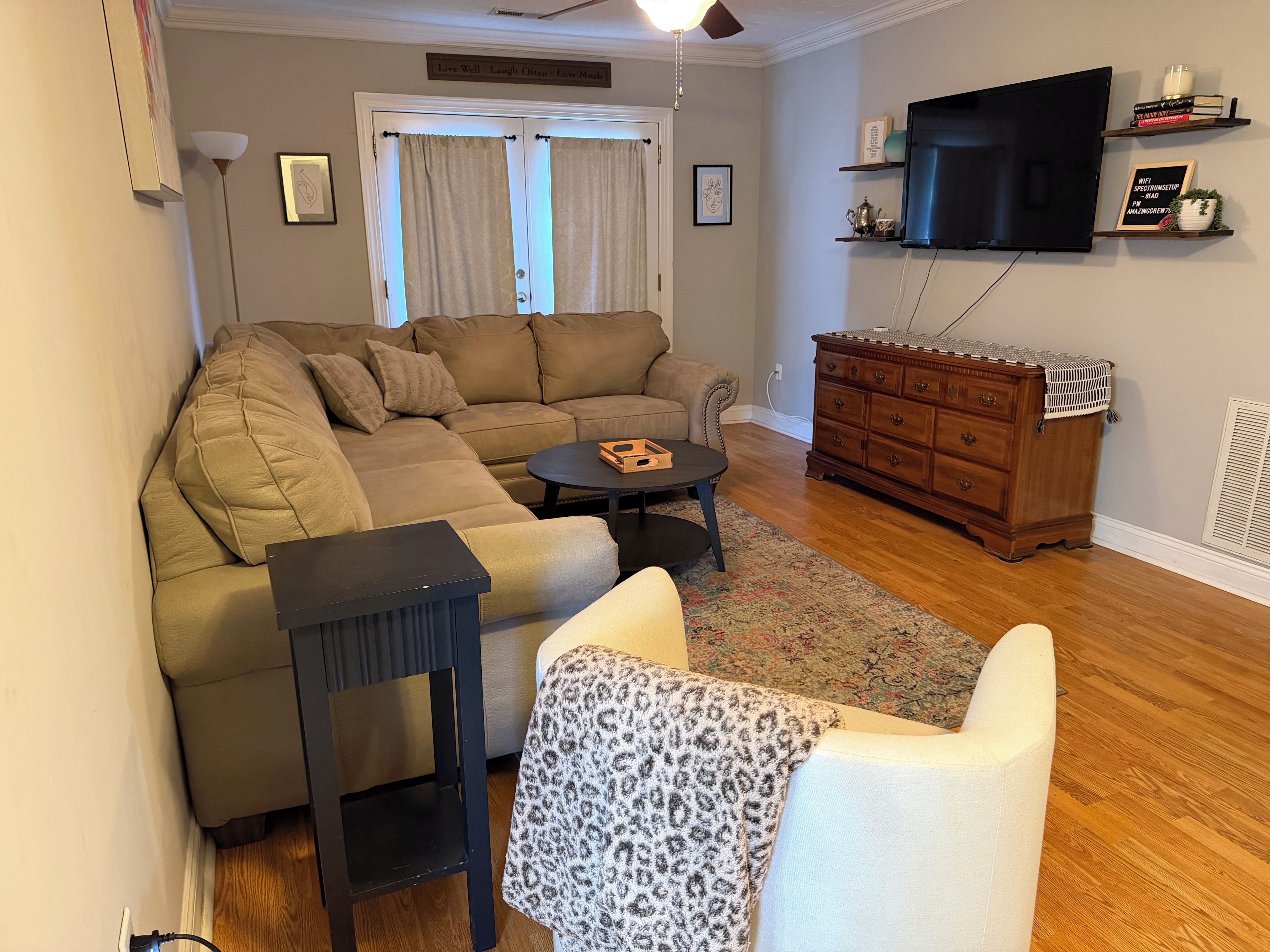 A living room with a sectional sofa, a coffee table, a wooden dresser, and a flat-screen TV mounted on the wall.