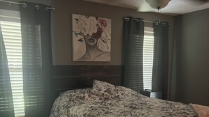 A bedroom with a bed, dark curtains, a ceiling fan, and a floral painting on the wall.