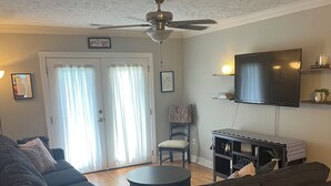 A living room with a dark-colored sofa, a round coffee table, a television mounted on the wall, and a ceiling fan.