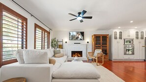 A modern living room with a white sofa, a fireplace, a ceiling fan, and a kitchen with white cabinets. - Coachella Casa Spanish-Style Oasis with Pool in Indio (Indio)