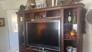 Una sala de estar con un televisor de pantalla plana grande, un mueble de madera para entretenimiento, un reposapiés de cuero y varios objetos decorativos.