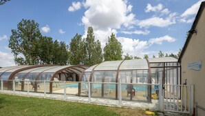 A covered swimming pool with a glass enclosure, a poolside area with lounge chairs, and a building with a sign. - Mobile Home in Veuzain-sur-loire With Garden (Veuzain-sur-Loire)