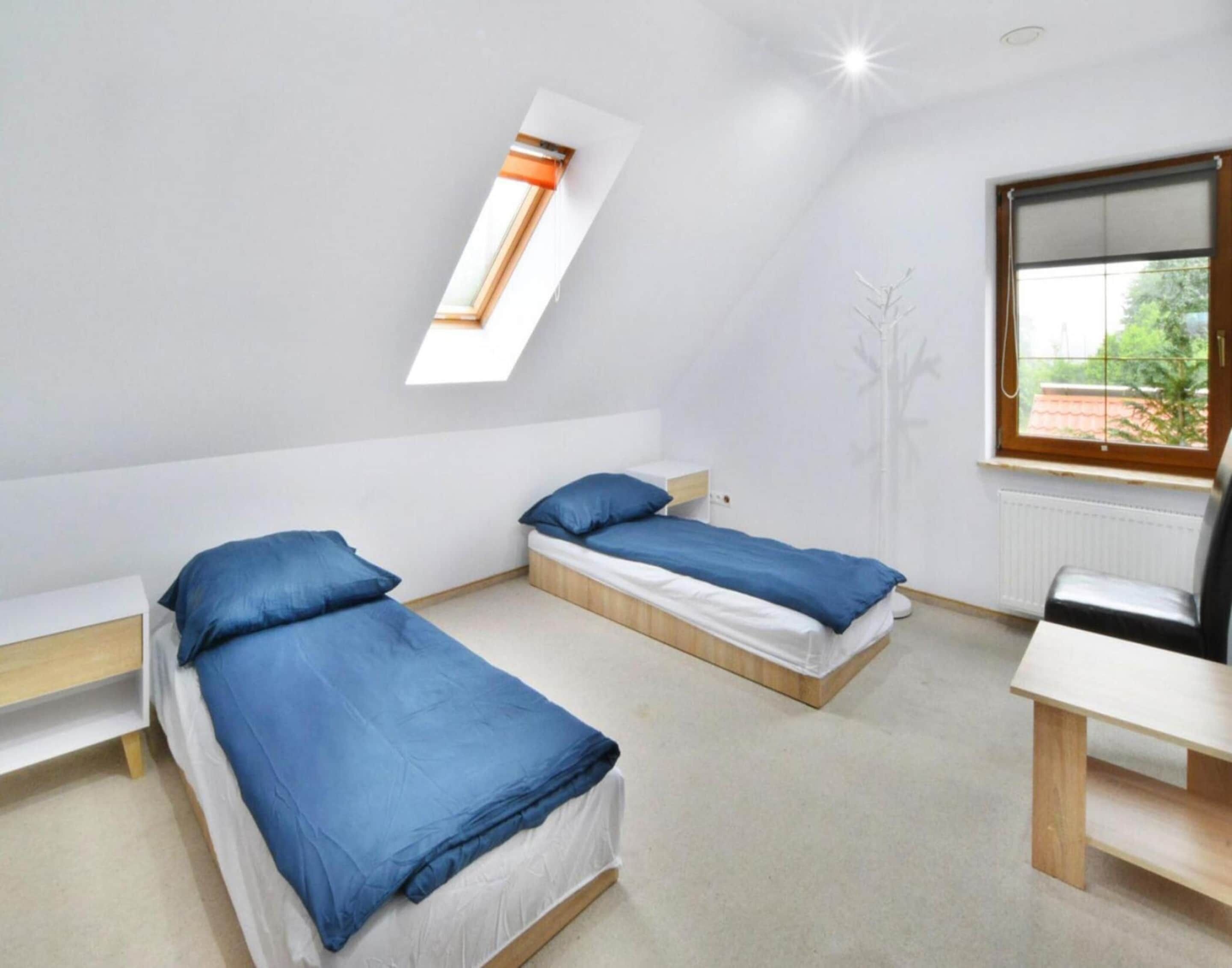 A dormitory room with two single beds, a desk, a chair, and a window with a view of greenery.