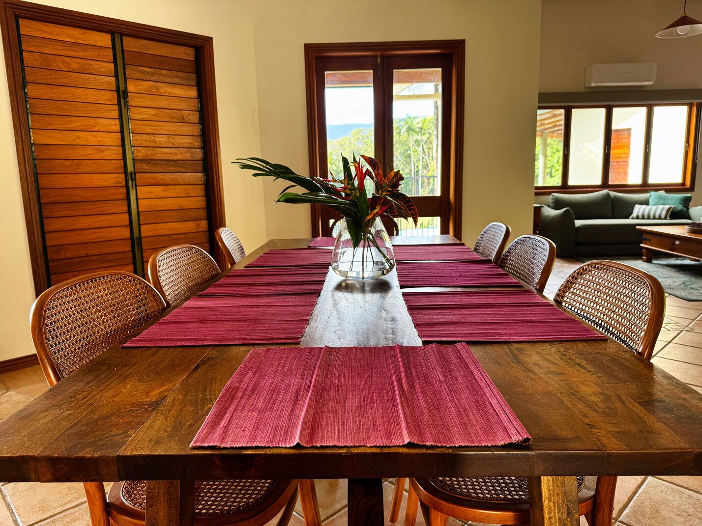 Una mesa de comedor con un florero, rodeada de sillas de mimbre, en una habitación con puertas de madera y vistas al exterior.