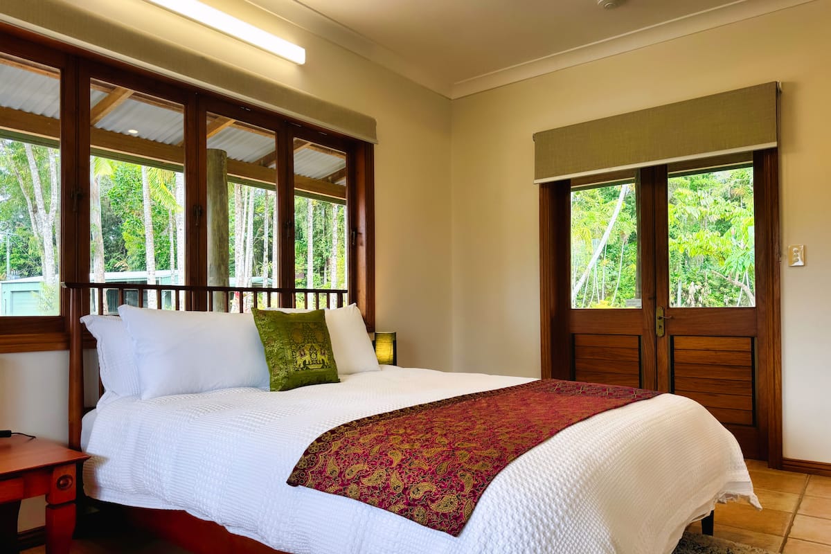 A bedroom with a large bed, wooden headboard, and a view of lush greenery through the windows.