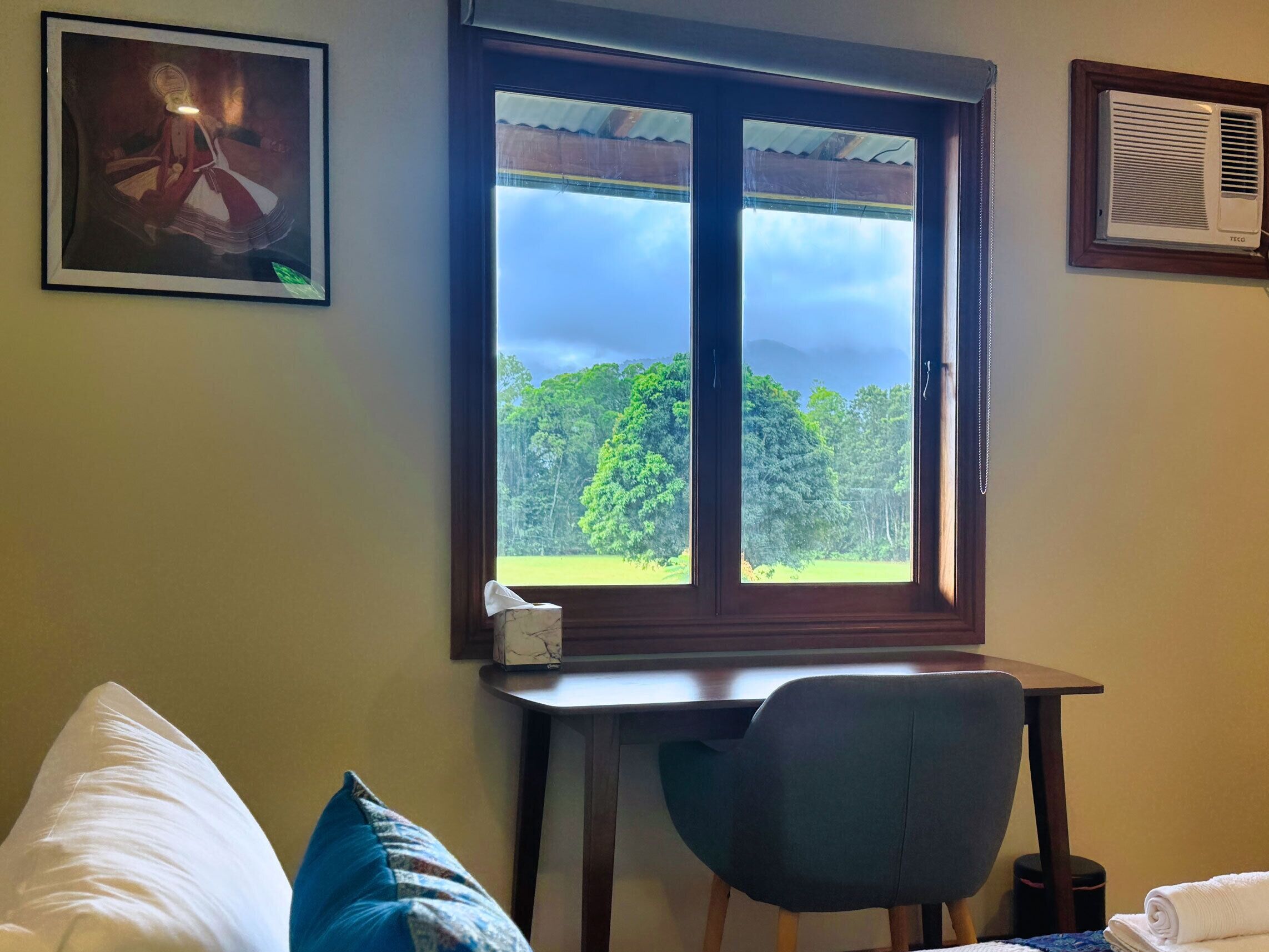 A room with a large window, a desk, a chair, and a bed with pillows.