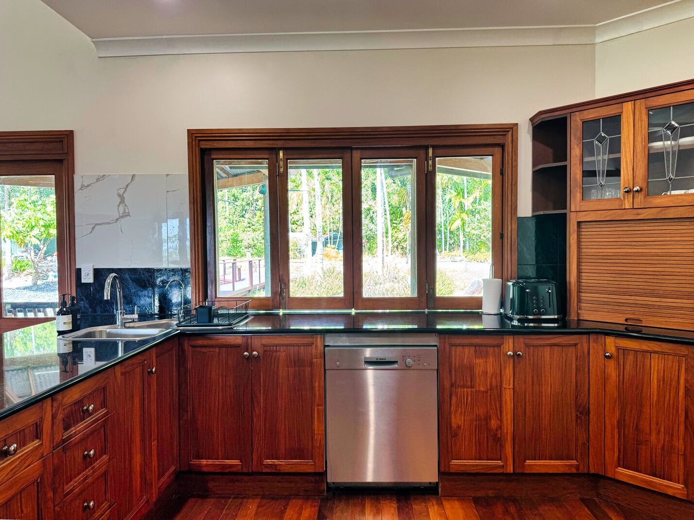 A modern kitchen with wooden cabinets, a dishwasher, and a large window overlooking a garden.