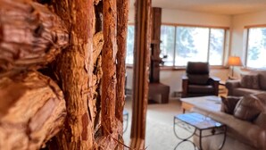 Een rustiek interieur met een houten wand, een zithoek met bank en salontafel, en een raam met uitzicht op bomen.