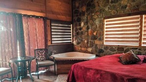 Una habitación con pared de piedra, techo de madera, colcha roja, una silla, una mesita y una bañera.