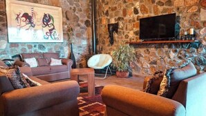 Una sala de estar acogedora con pared de piedra, un sofá marrón, una silla blanca y un televisor de pantalla plana sobre una estantería.