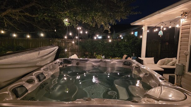 Ein Whirlpool mit Holzterrasse und Lichterkette, umgeben von einem Holzzaun und Bäumen.