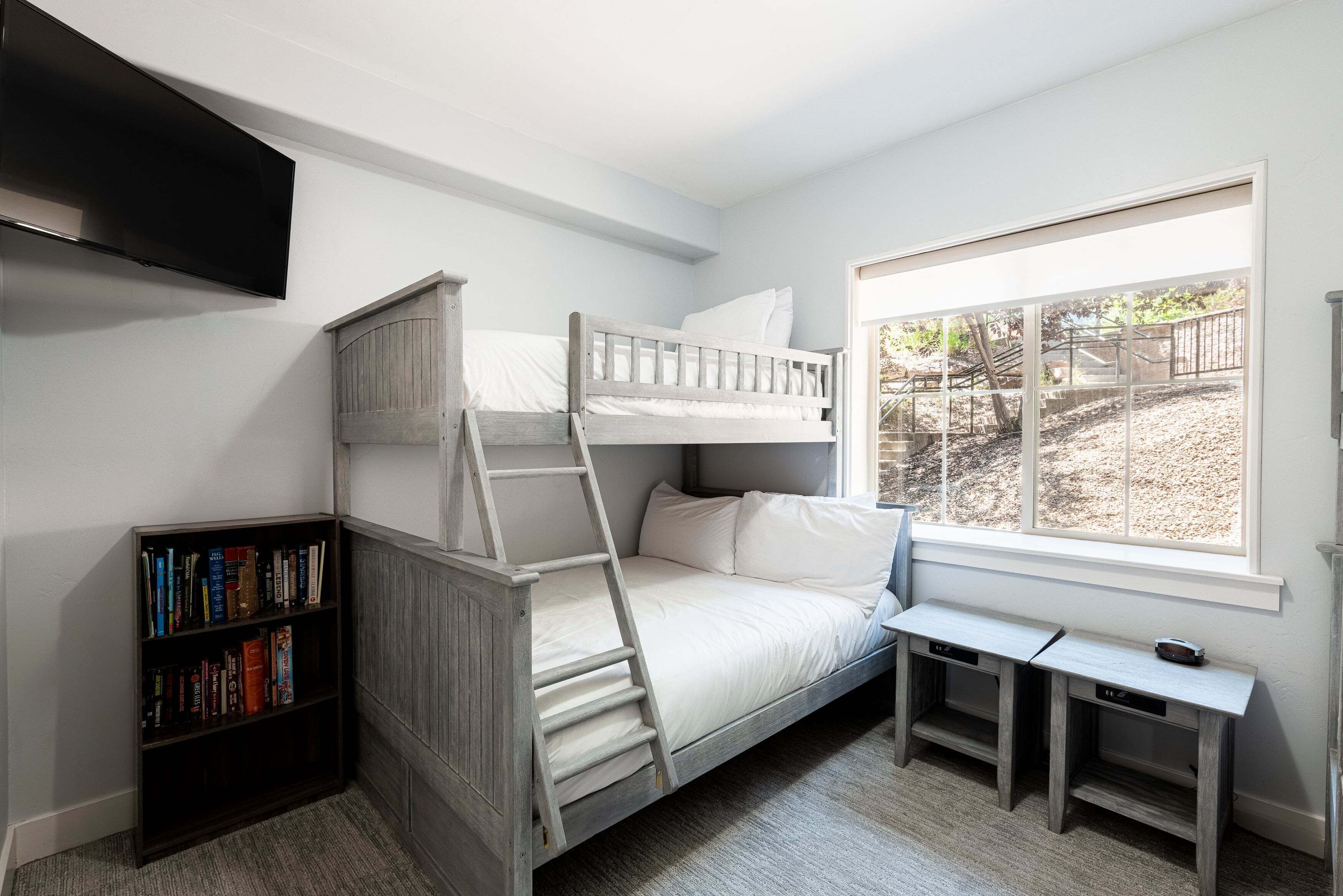 A bunk bed with a ladder, a bookshelf, a desk, and a window with a view of a garden.