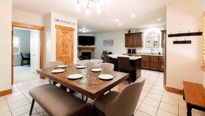 Un coin repas avec une table en bois dressée pour six personnes, un téléviseur en arrière-plan et une cuisine dotée de meubles en bois et d’un sol carrelé.