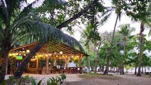 Ein Restaurant direkt am Strand mit einem Reetdach, Terrassenplätzen und Lichterketten.