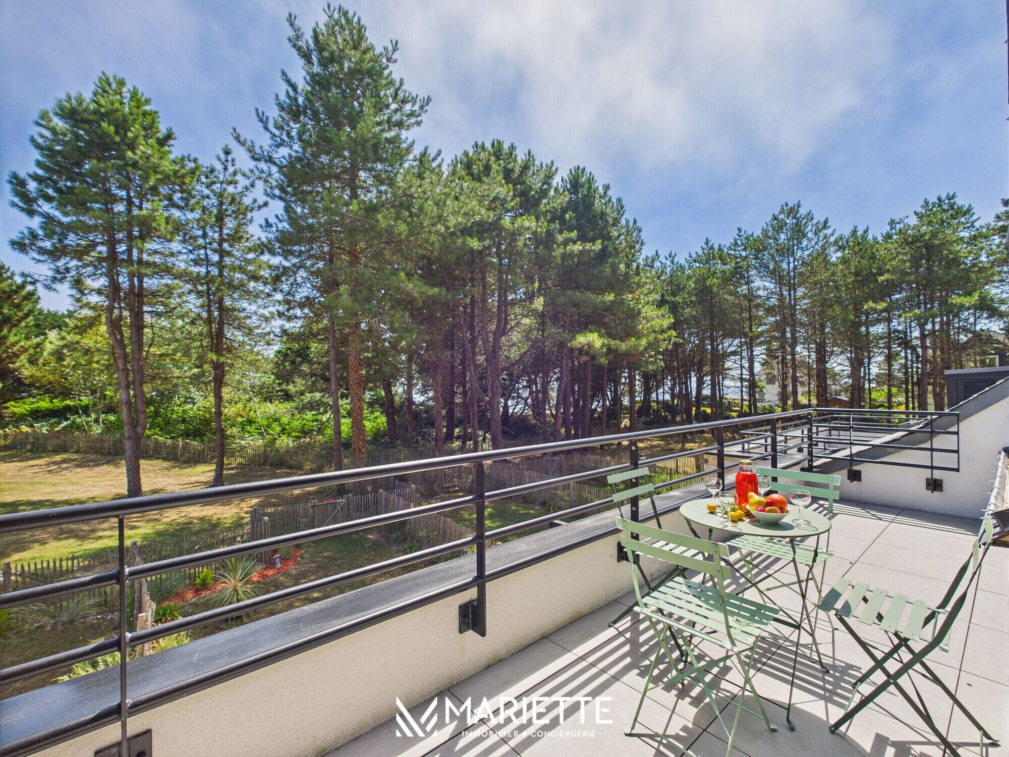A balcony with a view of a forest, a table with a fruit bowl, and a metal railing.