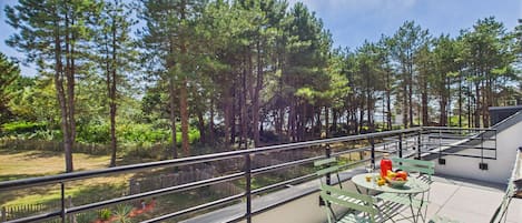 Uma varanda com vista para uma floresta, uma mesa com uma cesta de frutas e um corrimão de metal.