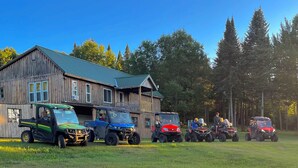A rustic wooden building with a green roof, surrounded by a grassy field and tall trees. - Escape to the Woods! Farmhouse on 10 acres -Great Wifi, Big Kitchen. BOOK IT! (Saint Regis Falls)