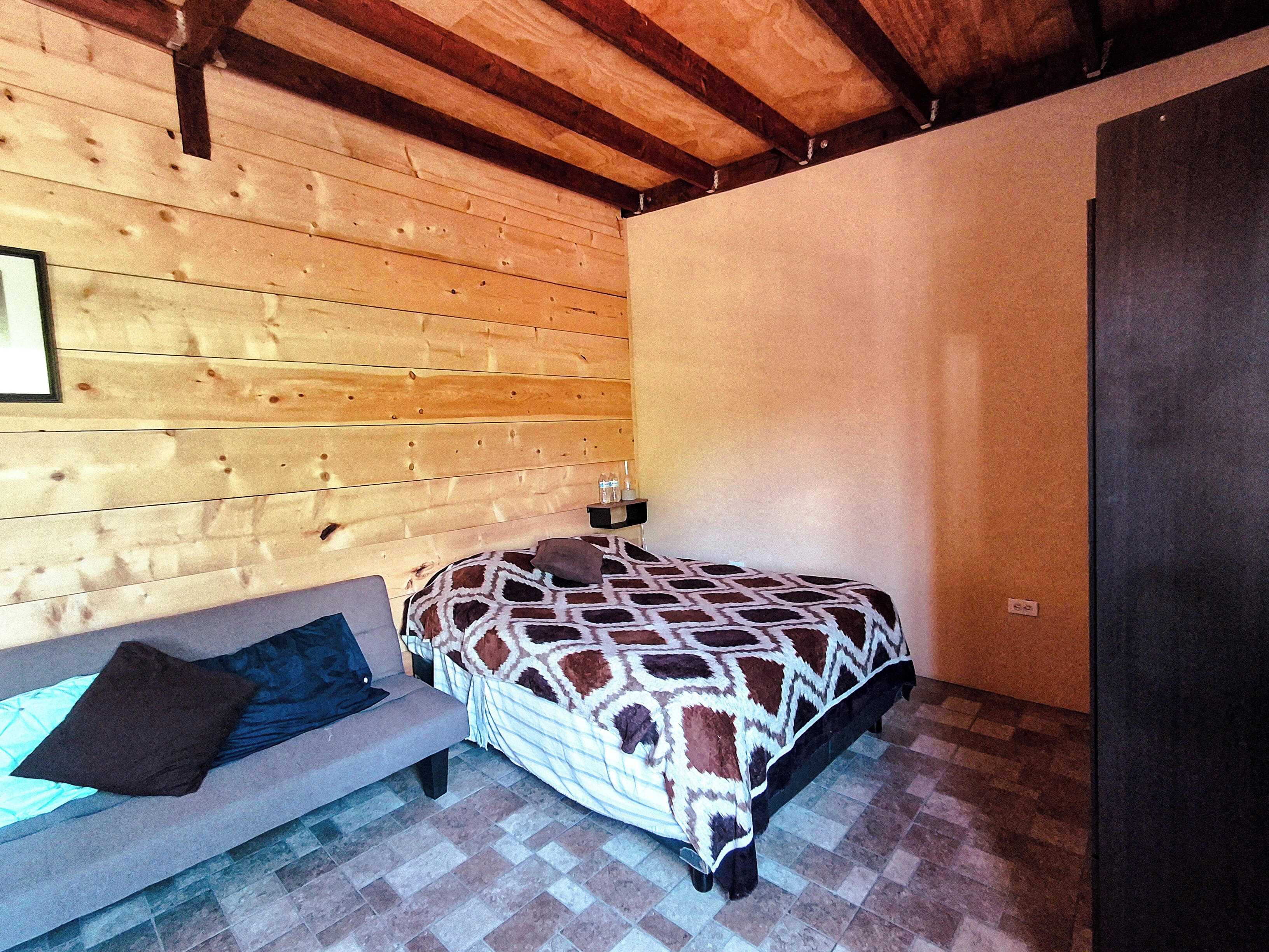 Una habitación con paneles de madera, una cama, un banco y una mesita de noche con botellas.