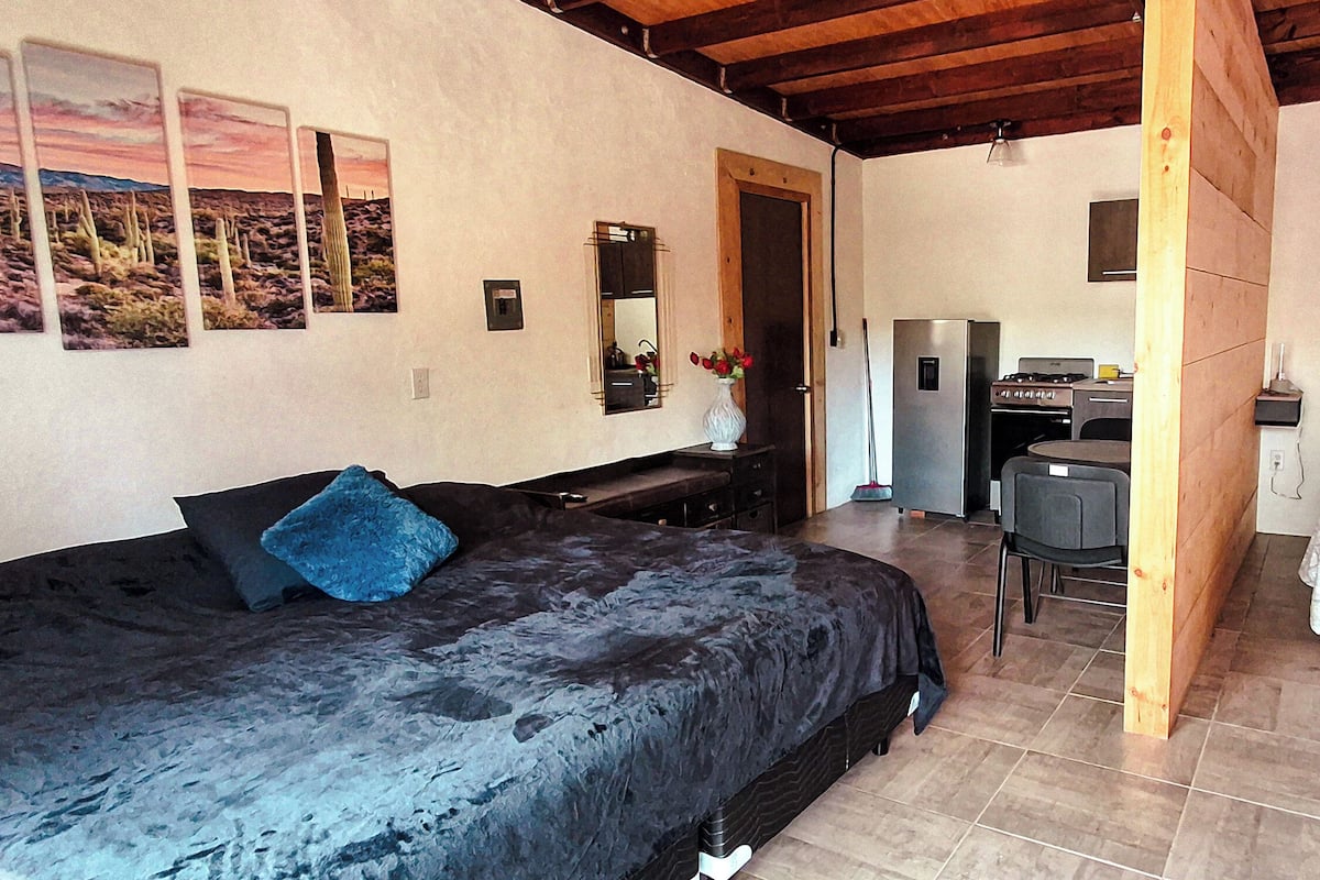 A bedroom with a bed, a desk, and a chair. There is a refrigerator and a mirror on the wall.
