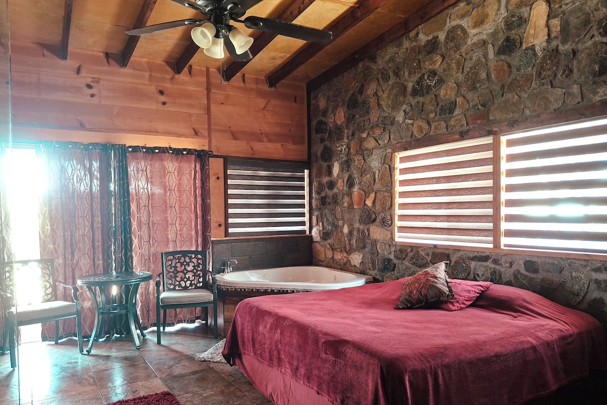 A bedroom with a stone wall, a wooden ceiling, a bed with a red bedspread, a chair, a small table, and a bathtub.