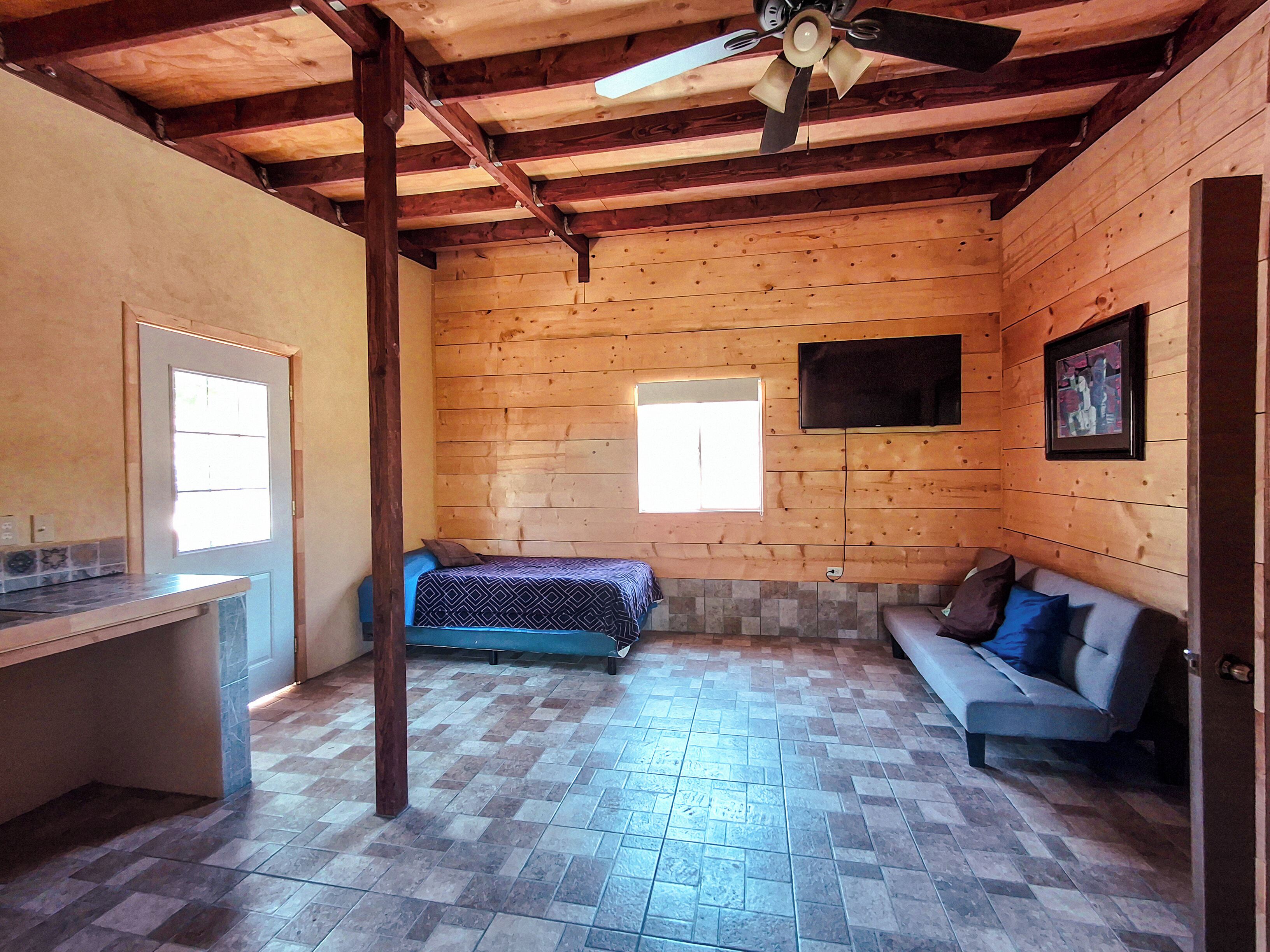 A room with a wooden ceiling, a bed, a sofa, a TV, and a window.