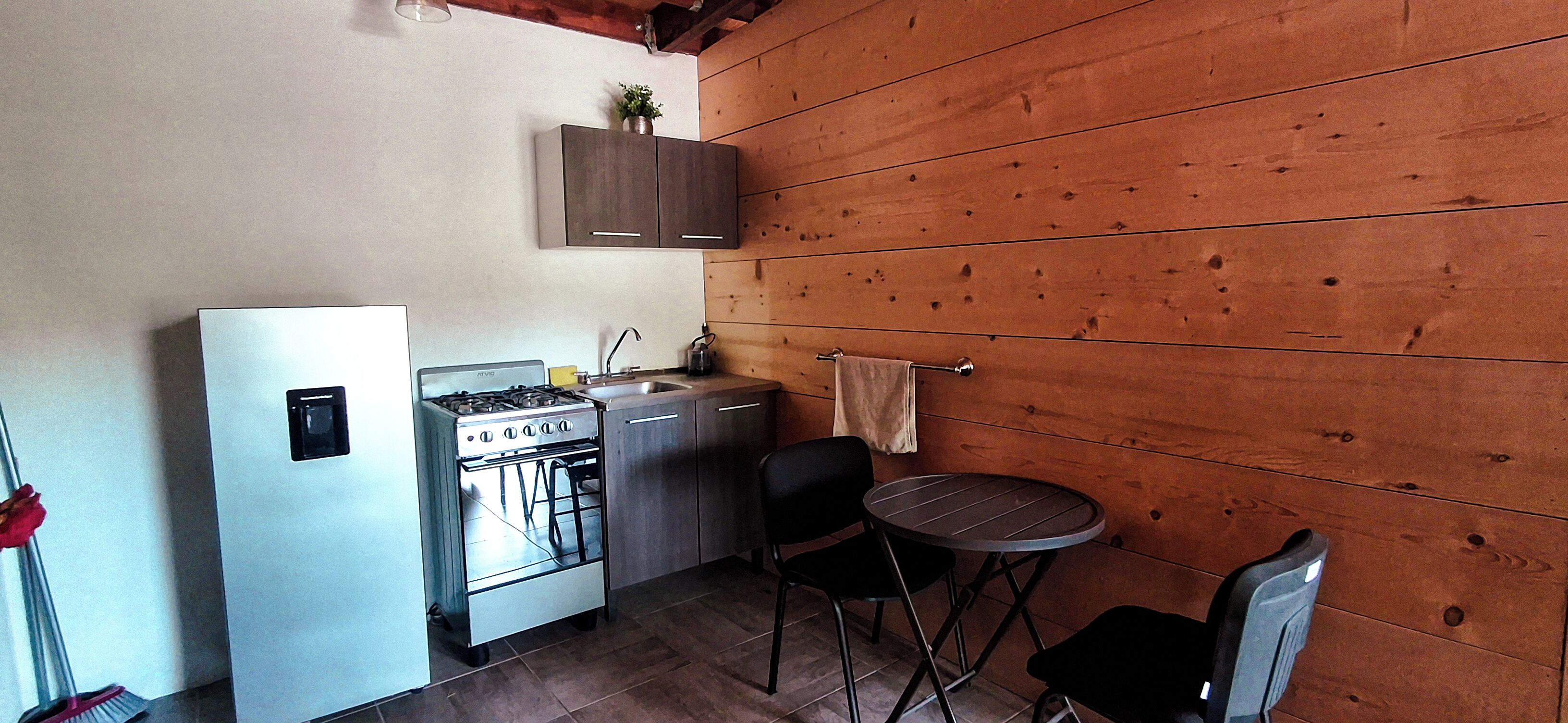 Una cocina compacta con estufa, lavamanos y armarios, una pequeña mesa con sillas, y una pared de madera.
