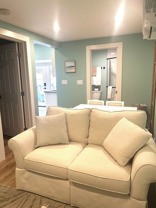 A beige sectional sofa with a light-colored rug, a white door, and a bathroom visible through an open door.