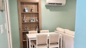 Un petit coin repas avec une porte en bois, une table et des chaises blanches, un canapé blanc et un climatiseur fixé au mur.