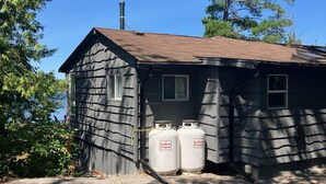 A small, single-story building with a sloped roof and a chimney. In front of the building, there are two propane tanks labeled "Indecott Propane." The building is surrounded by trees and gravel.