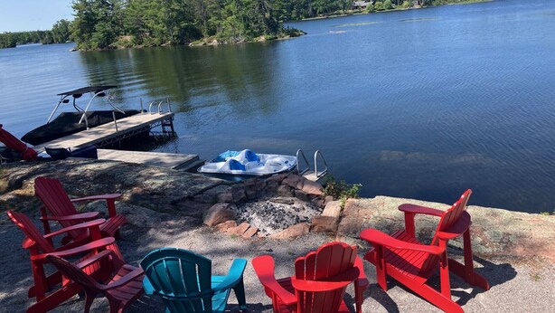 A lakeside setting with a dock, a small boat, and red and blue Adirondack chairs.