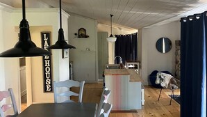 A dining area with a long table and chairs, a kitchen in the background, and a ceiling with exposed wooden beams.