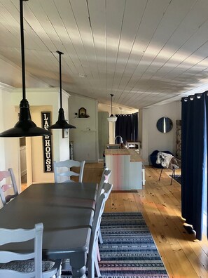 Uma sala de jantar com uma mesa comprida e cadeiras, uma cozinha ao fundo e um teto com vigas de madeira aparente.