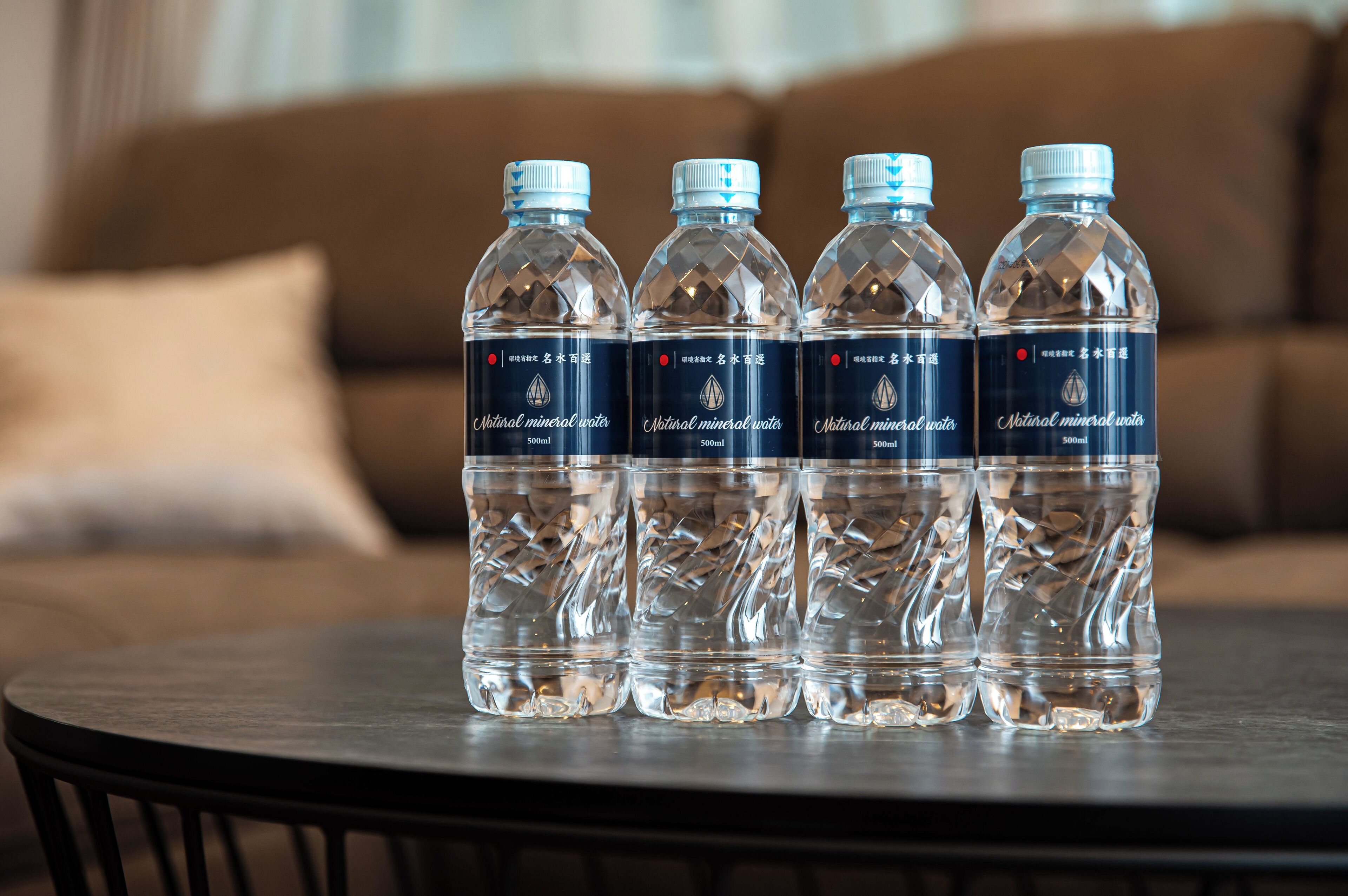 Vier flessen water op een ronde tafel, met een vage achtergrond van een bank en kussens.