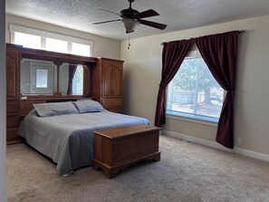 A bedroom with a bed, wooden wardrobe, a chest of drawers, a ceiling fan, and a window with curtains.