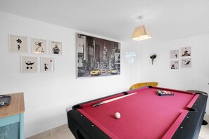 Ein Billardtisch mit roter Spielvliesdecke, umgeben von gerahmten Kunstwerken an den Wänden, und ein Neonzeichen.