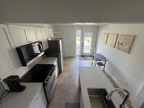 A compact kitchen with white cabinets, a built-in microwave, a stove, a refrigerator, and a small dining area with a table and chairs. - 1-Large bedroom Suite in quiet neighborhood. (Vernon)