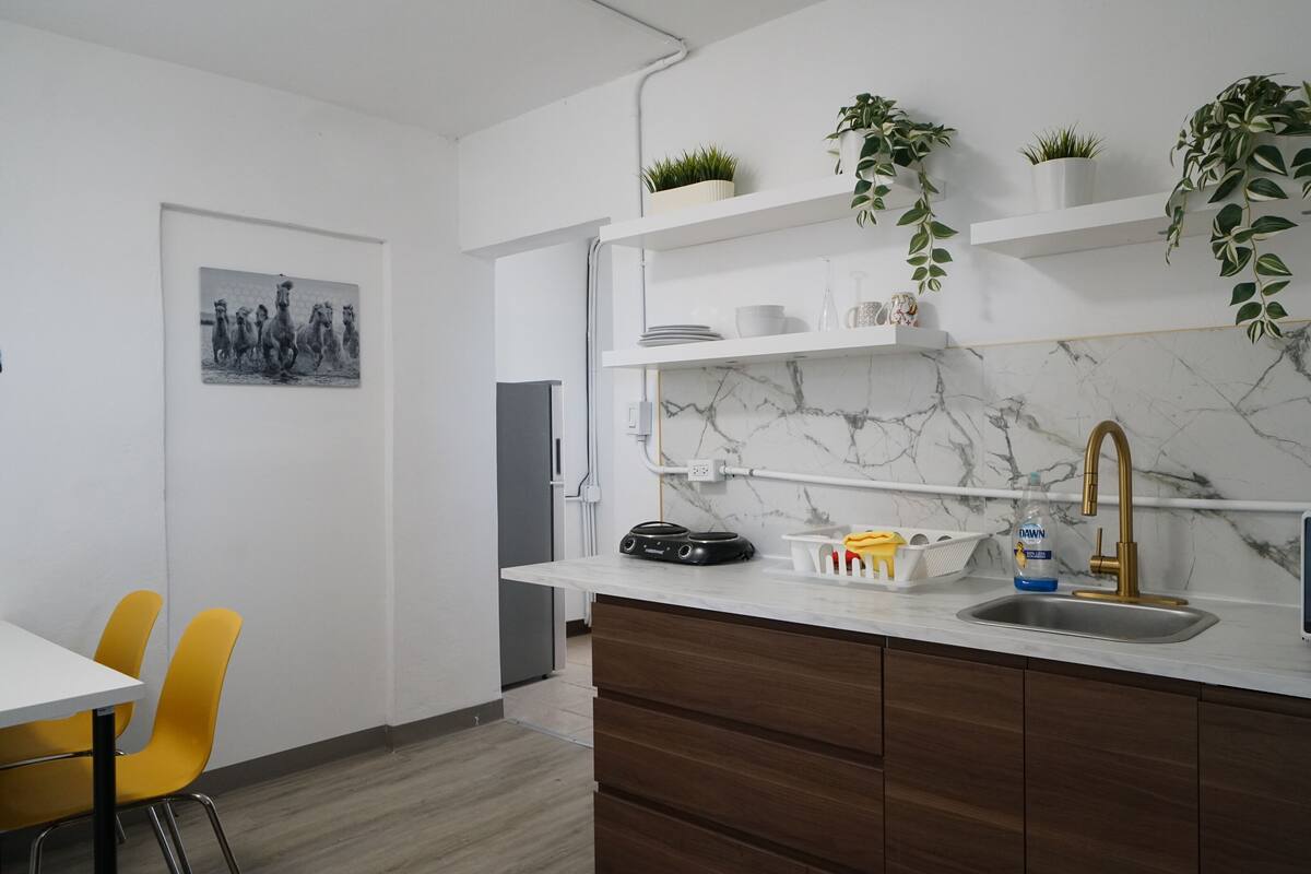 A modern kitchen with a marble backsplash, a built-in dishwasher, and a black refrigerator. There are yellow chairs and a dining table visible.