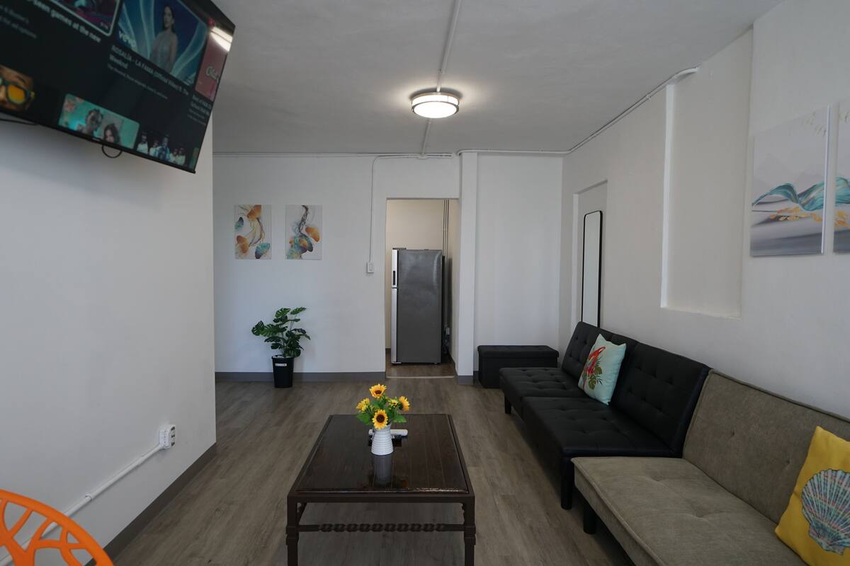 A modern living room with a sofa, a coffee table with a vase of sunflowers, a potted plant, and a flat-screen TV mounted on the wall.