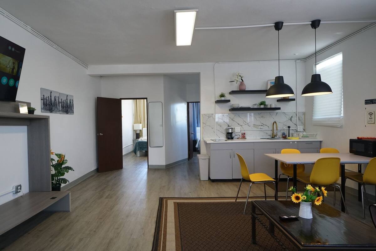 A modern kitchen with a built-in microwave, a dining area with yellow chairs, and a living room with a flat-screen TV.