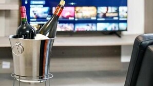A modern living room with a black leather sofa, a small white table with a metal container holding a bottle and a champagne flute, and a large flat-screen TV displaying a sports channel. - CARIBBEAN STARS (Carolina)