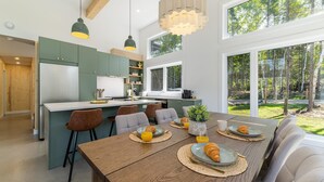 A modern kitchen with a central island, wooden dining table with chairs, and large windows offering a view of the outdoors.