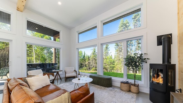 A spacious living room with a leather sofa, a round coffee table, and a fireplace. Large windows offer views of a forested area.