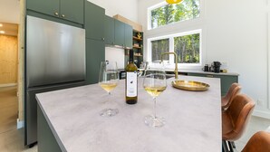 A modern kitchen with a large island, stainless steel appliances, and a view of the forest through the windows.